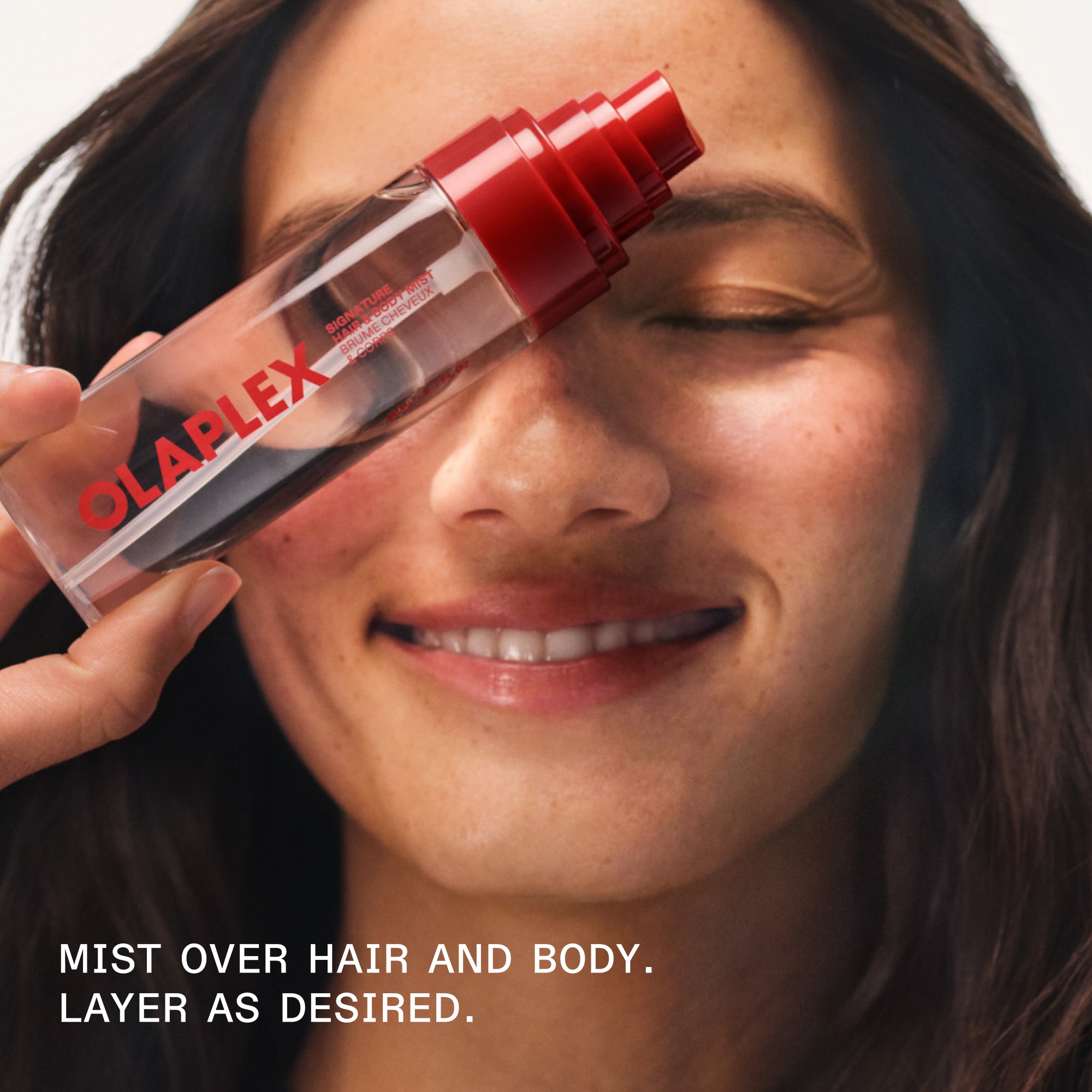 Une personne souriante tenant un flacon de brume pour cheveux et corps OLAPLEX. Le texte superposé indique : « MIST OVER HAIR AND BODY. LAYER AS DESIRED. » (Brumiser sur le corps et les cheveux. Superposer au gré des envies.).